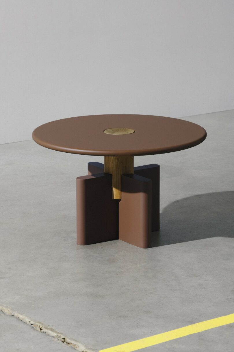 A round brown table with a central accent, supported by a geometric base, placed on a concrete floor near a yellow painted line.