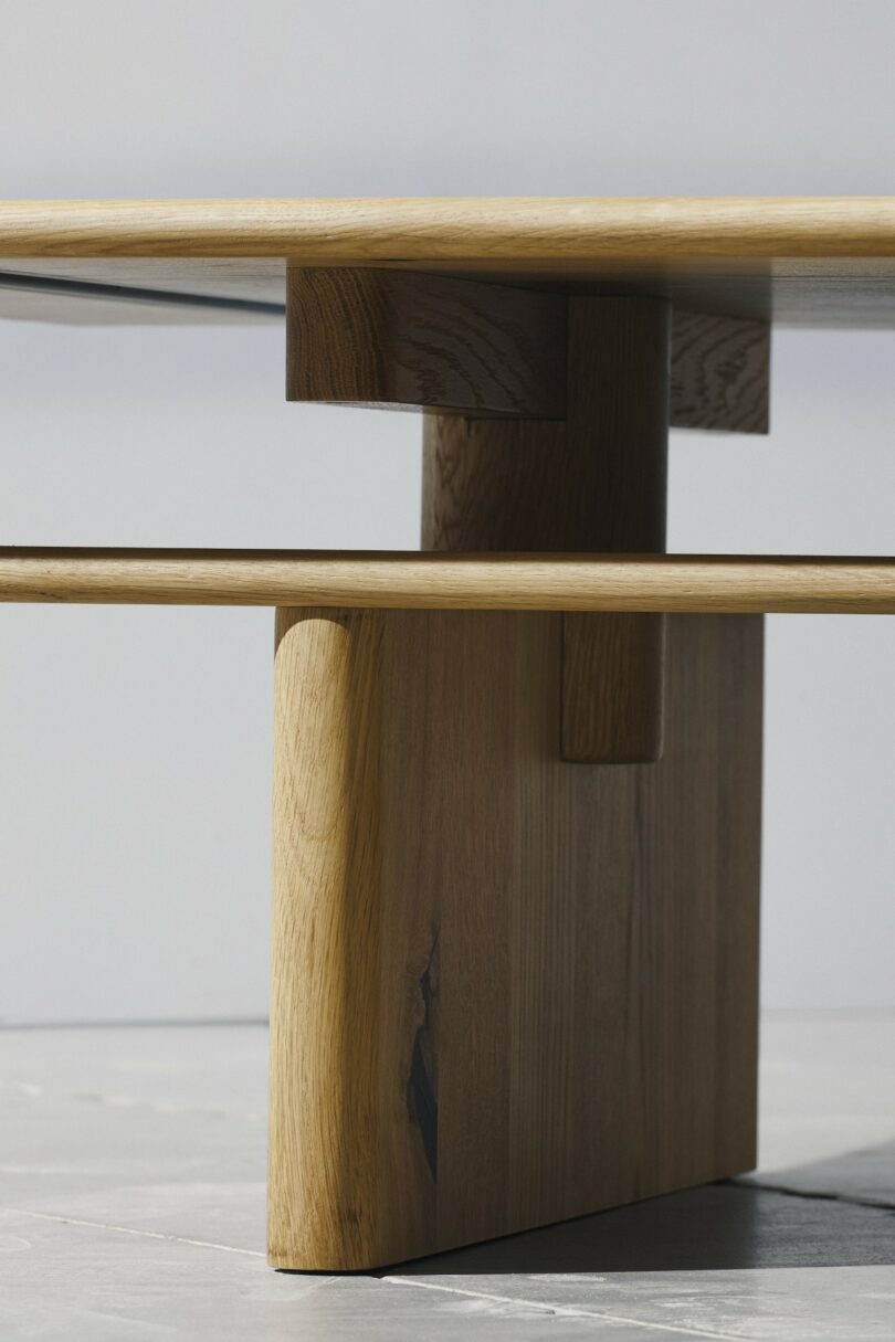 Close-up view of a modern wooden table with geometric lines and a natural finish, showing the underside and sturdy vertical support on a light floor