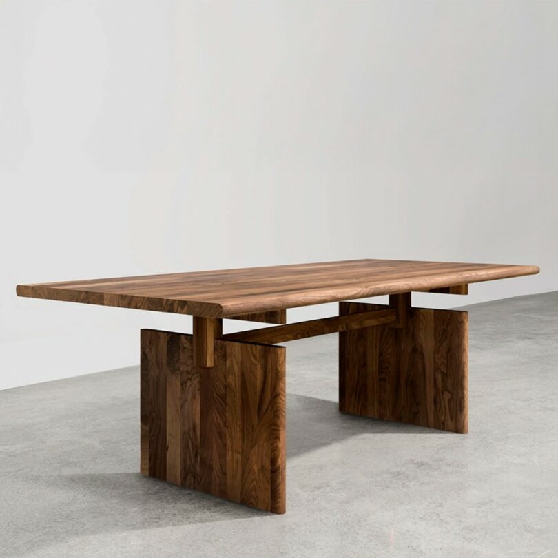 A modern rectangular wooden table with a thick top and two wide, flat legs, set against a plain gray background