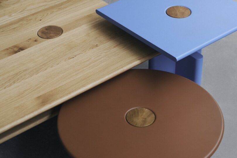 Three overlapping tabletops in wood, blue, and brown finishes, each featuring a circular wooden inlay at the corner or center, set against a neutral background