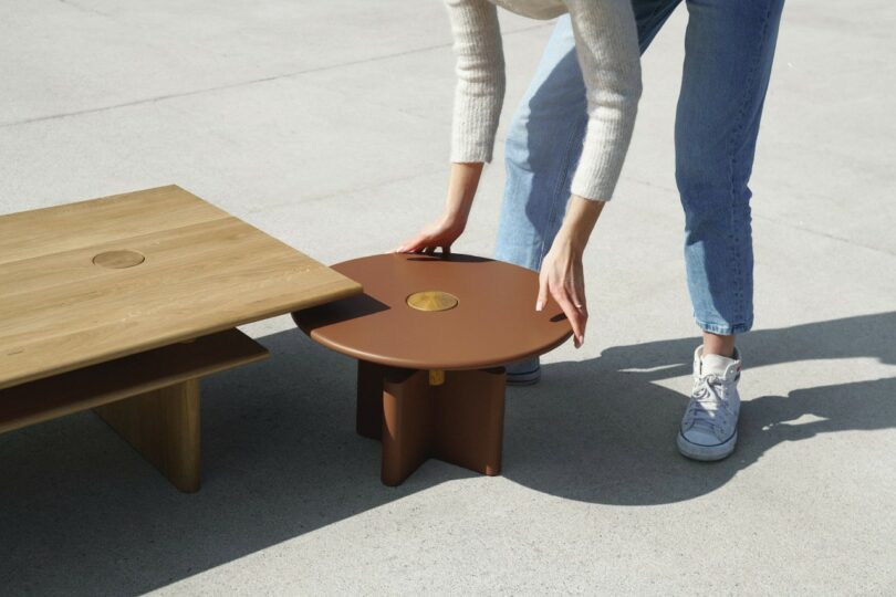 A person in jeans and sneakers adjusts a small round brown table next to a larger rectangular wooden table on a smooth surface.