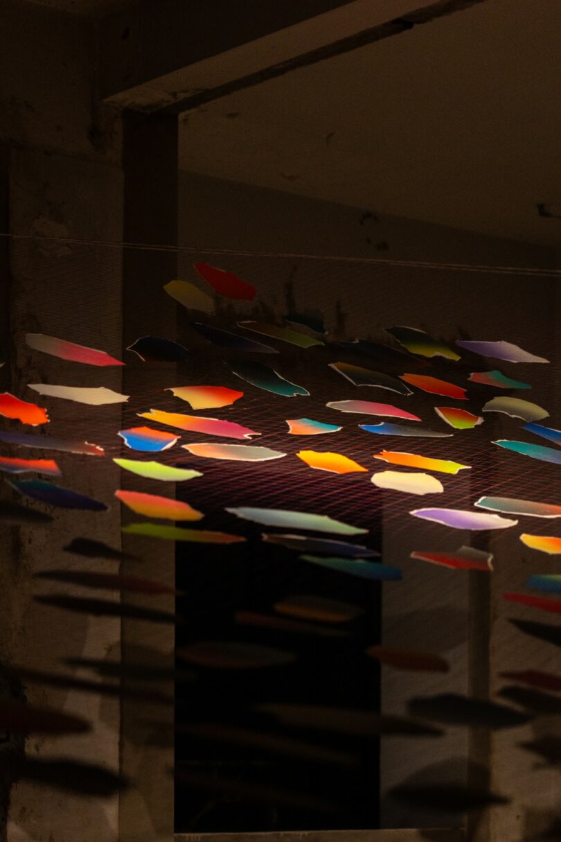 Colorful paper shapes hang from strings indoors, casting shadows on the wall and ceiling in a dimly lit room with exposed concrete beams