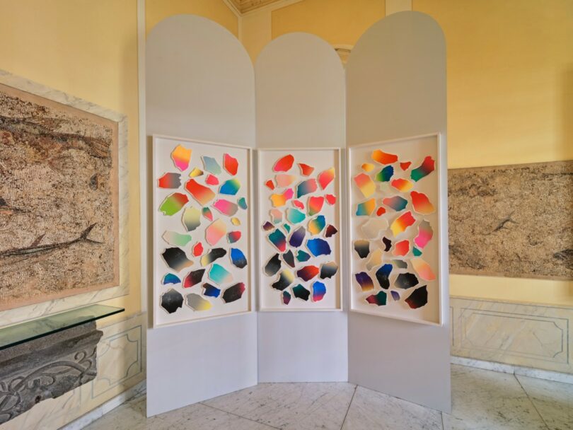 Three white folding panels display colorful abstract shapes in a bright room with classical wall art and marble flooring