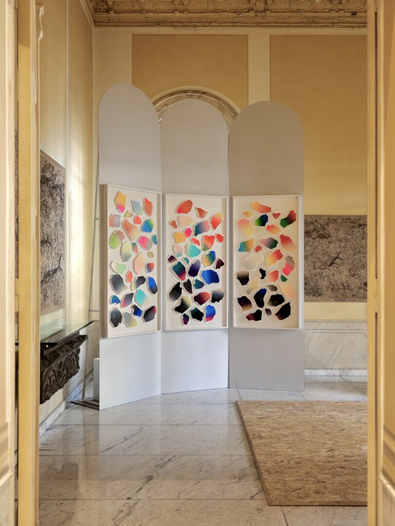 Three white folding panels display colorful abstract shapes in a bright room with classical wall art and marble flooring