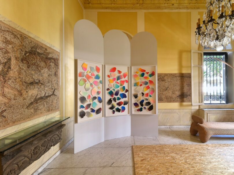 Three white folding panels display colorful abstract shapes in a bright room with classical wall art and marble flooring