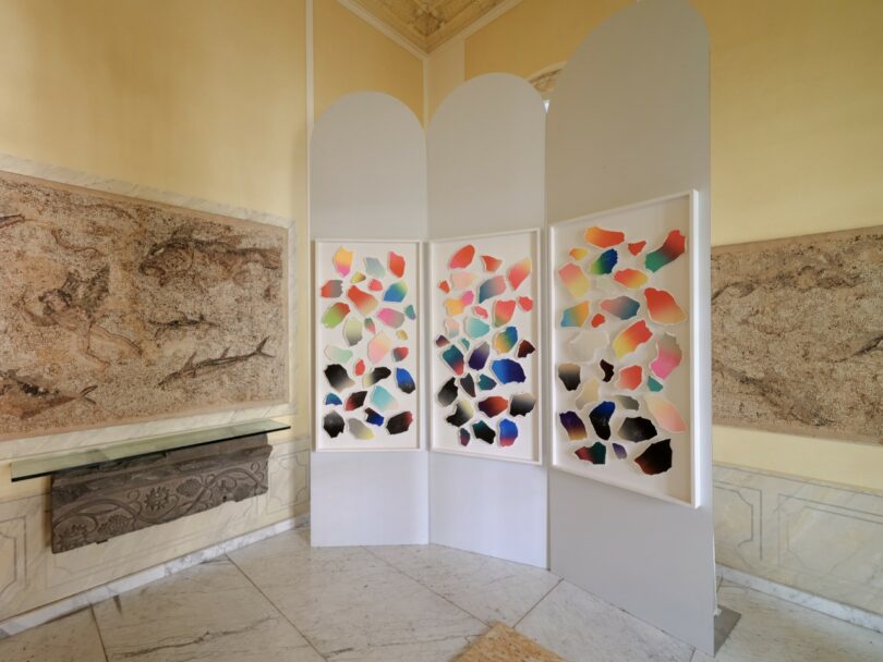 Three white folding panels display colorful abstract shapes in a bright room with classical wall art and marble flooring