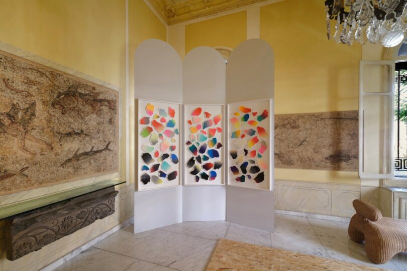 Three white folding panels display colorful abstract shapes in a bright room with classical wall art and marble flooring
