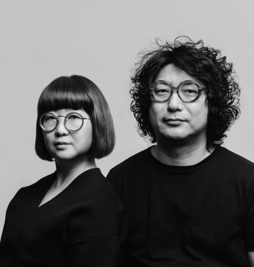 A woman and a man with round glasses and dark clothing pose together against a plain light background