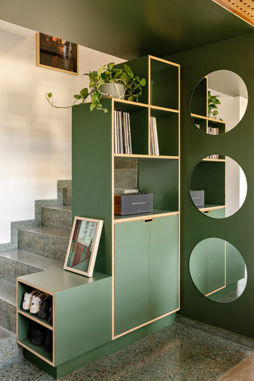 Modern green entryway storage unit with open shelves, a cabinet, a potted plant, three round mirrors, shoe cubbies, and a framed photo near stairs.
