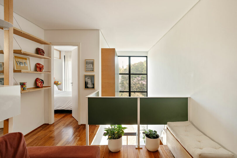 Modern upstairs hallway with wooden floors, green partition, potted plants, a built-in bench, shelving, and a view into a bright bedroom. Large window lets in natural light.
