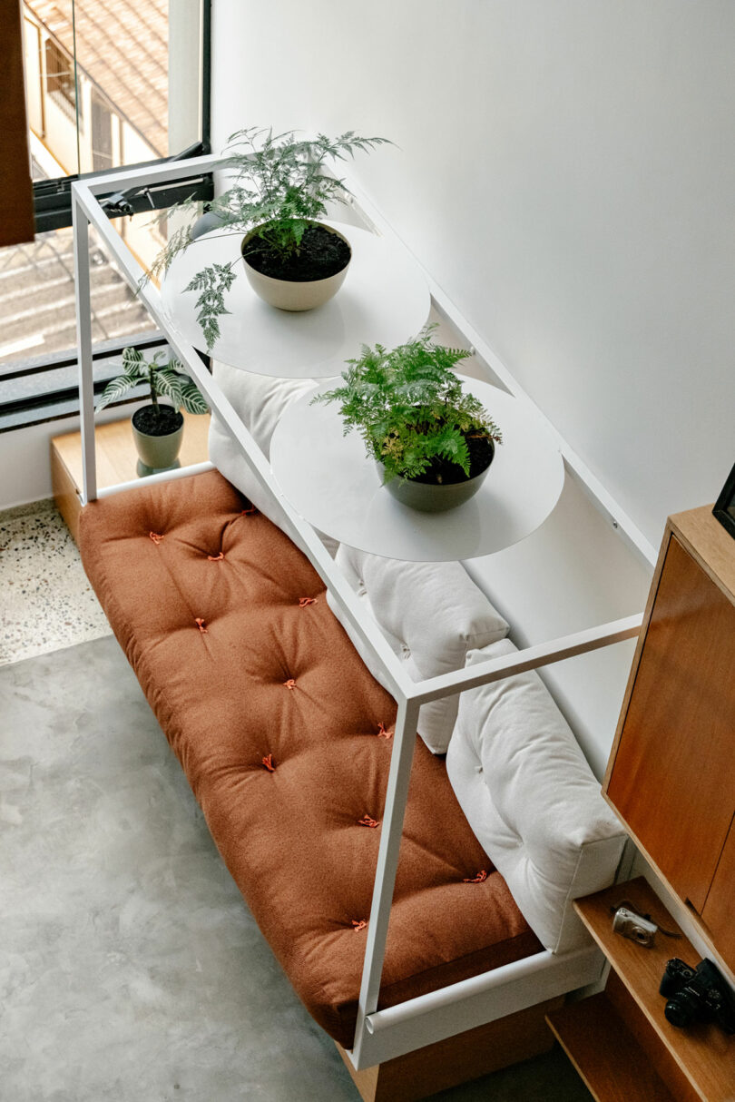 A modern indoor seating area with a brown cushioned bench, white pillows, and a white metal frame holding two potted plants on circular shelves above the bench.