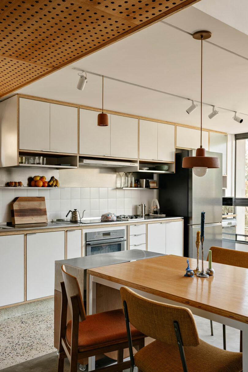 Modern kitchen with white cabinets, stainless steel appliances, wooden dining table, upholstered chairs, and built-in open shelves with dishes and fruit.