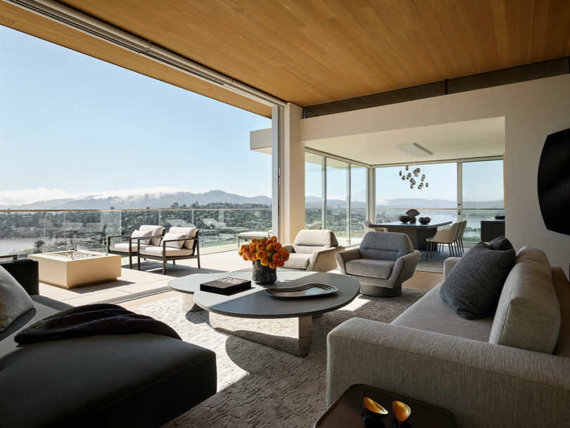 Modern living room with large windows, neutral furnishings, and a round coffee table; expansive views of mountains and water visible outside.