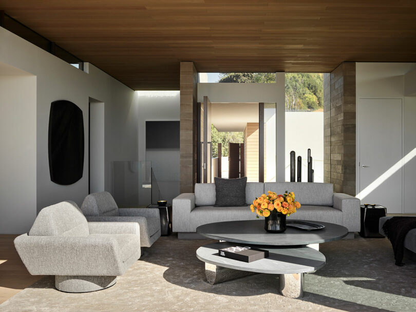 Modern living room with gray sofas, a round coffee table with flowers, wooden ceiling, large windows, and neutral decor.