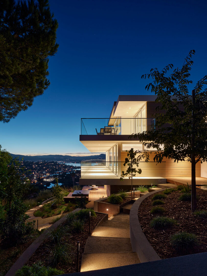 A modern house with large glass windows and balconies is illuminated at dusk, overlooking a cityscape and water in the distance, with landscaped gardens and pathways in the foreground.