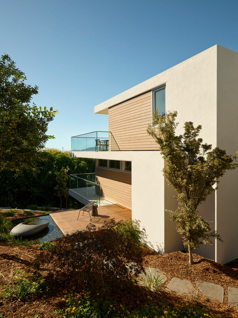 A modern two-story house with wood and white exterior walls, glass balconies, and a landscaped yard with trees, plants, and outdoor seating.