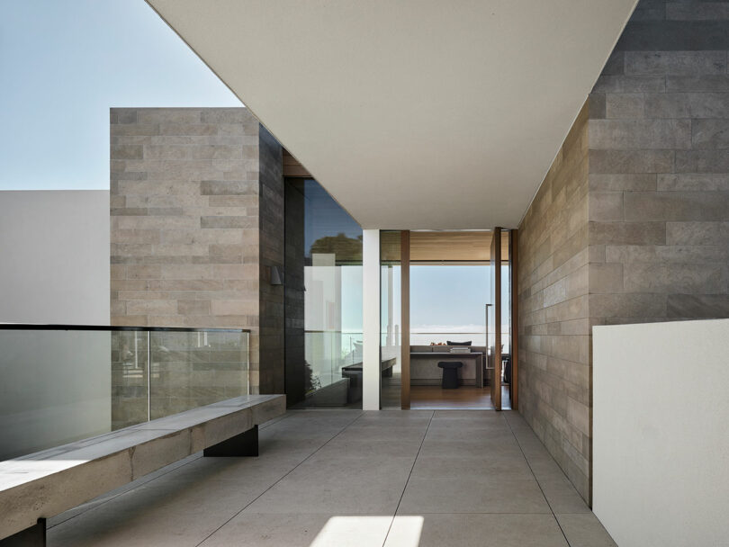 Modern building corridor with stone walls, glass railing, concrete bench, and view into a room with large windows and ocean in the background.