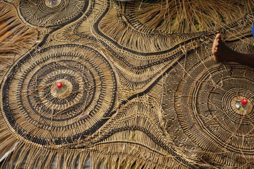 Intricately woven straw mat with circular patterns and two small red objects placed near the center of two circles; a partial view of a person's foot is visible on the right