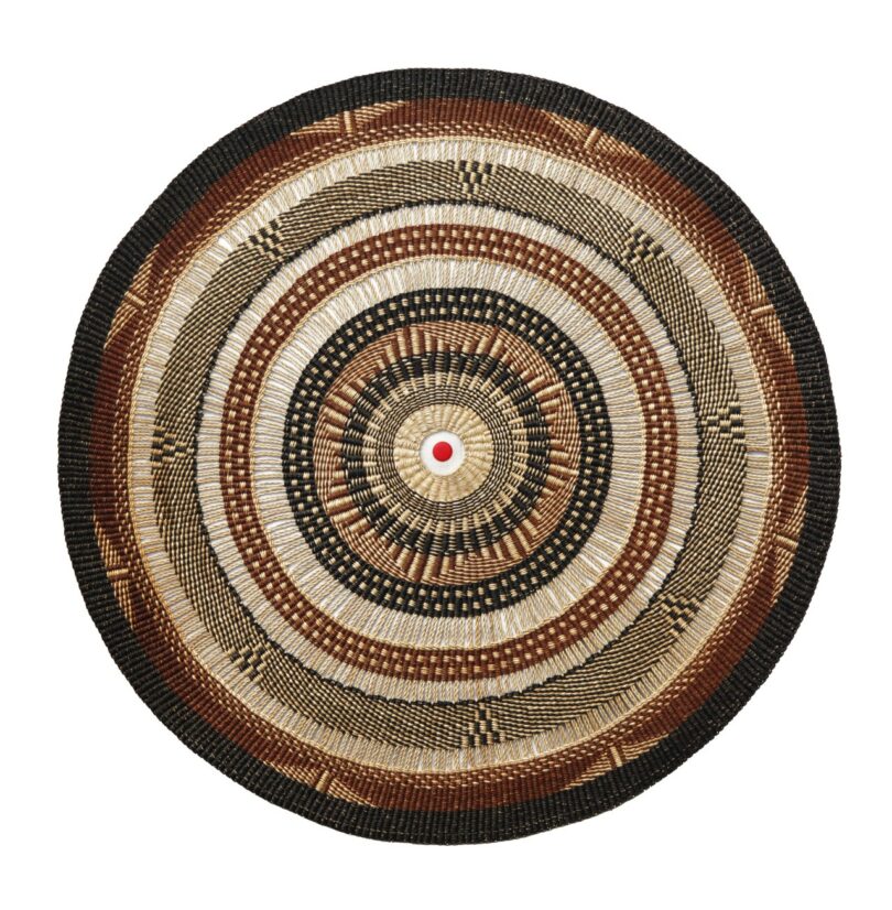 A round woven tapestry-lamp featuring concentric circles in black, brown, tan, and beige with a small red bottle cap in the center.