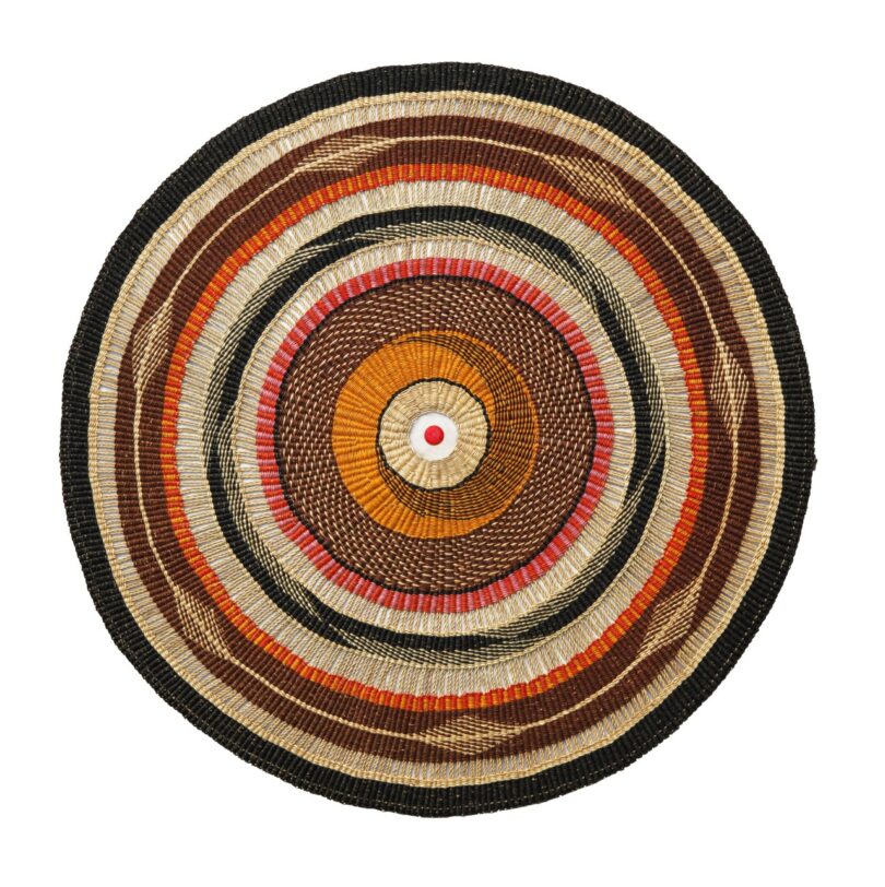 A round woven tapestry-lamp featuring concentric circles in black, brown, tan, and beige with a small red bottle cap in the center.