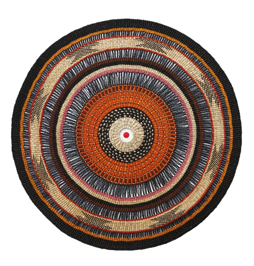 A round woven tapestry-lamp featuring concentric circles in black, brown, tan, and beige with a small red bottle cap in the center.