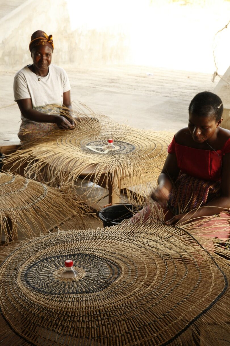 Two women sit on the floor weaving large, round baskets from natural fibers, each basket anchored at the center with a red bottle cap