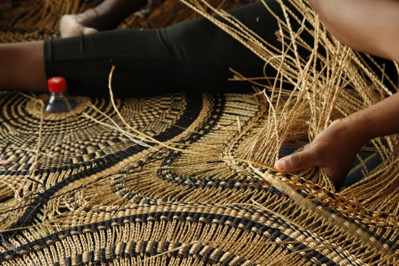 A person weaves an intricate basket using natural fibers, with their hands actively working and a patterned mat beneath. A red bottle cap is visible nearby