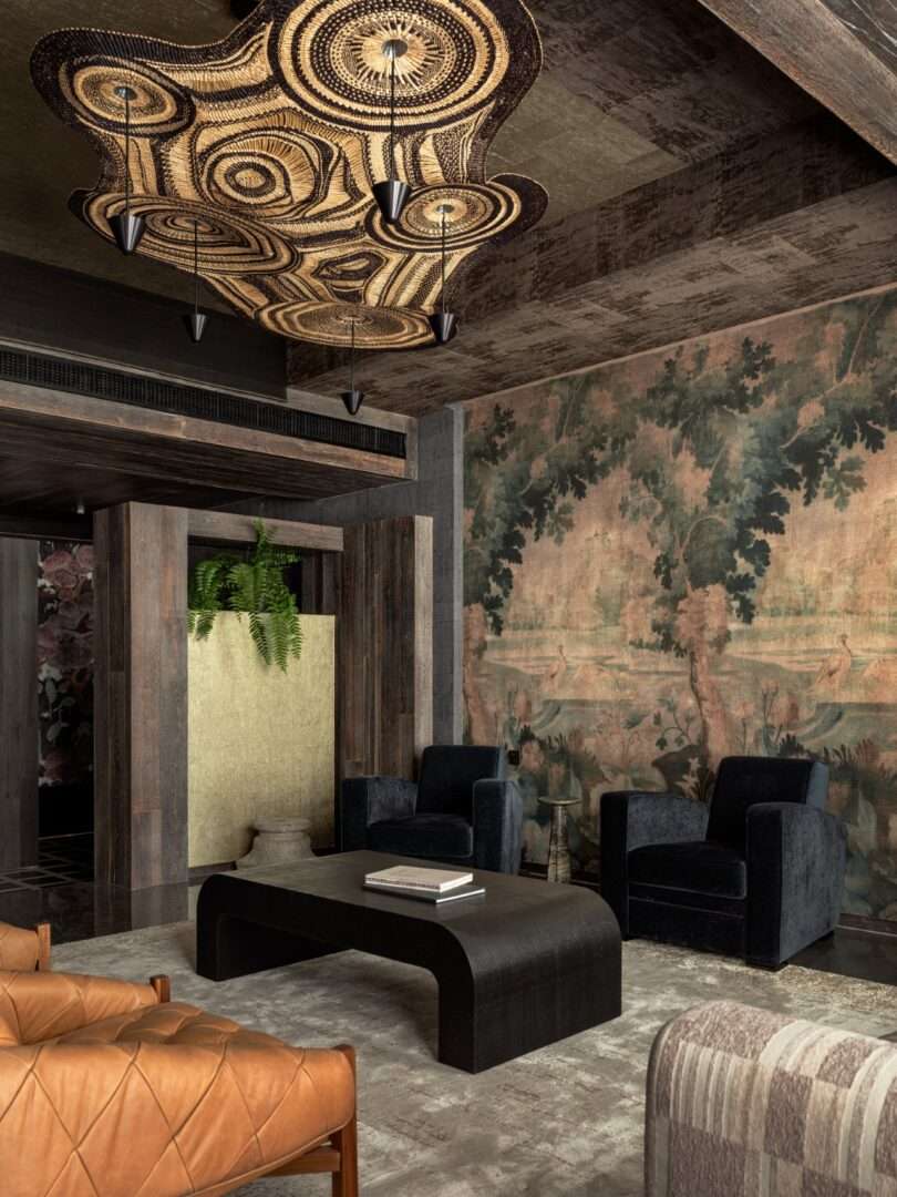 Modern living room with black armchairs, a curved black coffee table, tan leather seating, large abstract ceiling light, a wall mural of trees, and a hanging potted plant