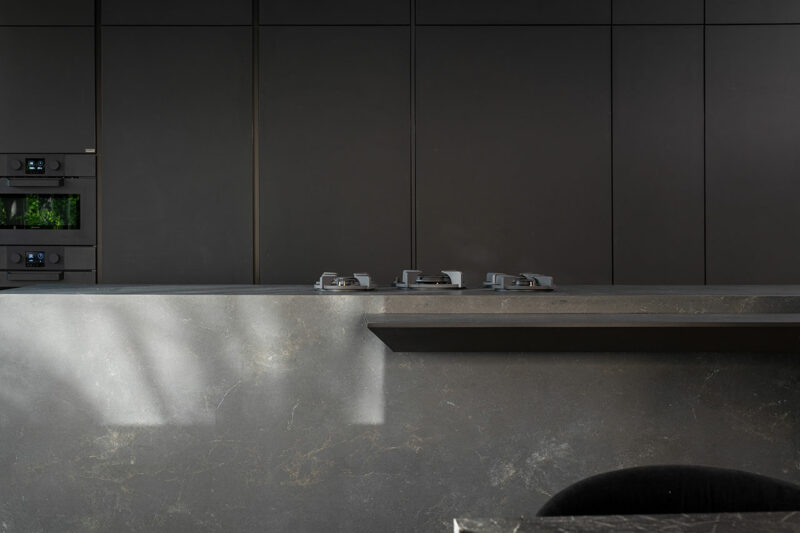 Modern minimalist kitchen with dark matte cabinetry, a built-in double oven, a sleek stone countertop, and three gas burners on the island.