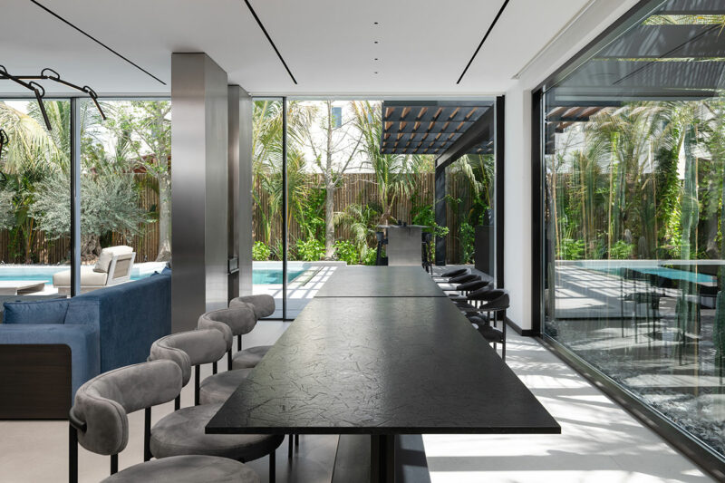 A modern dining area with a long black table and gray chairs, large windows overlooking a pool and garden, and an adjacent living room.