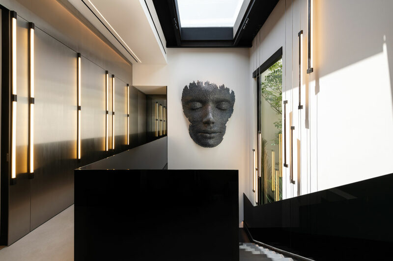 A modern interior with a black staircase, wall-mounted linear lights, and a large sculpted face artwork displayed on a white wall under a skylight.