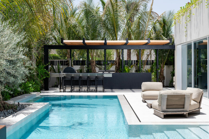Modern backyard with a swimming pool, poolside lounge chairs, and an outdoor kitchen area under a pergola surrounded by palm trees.