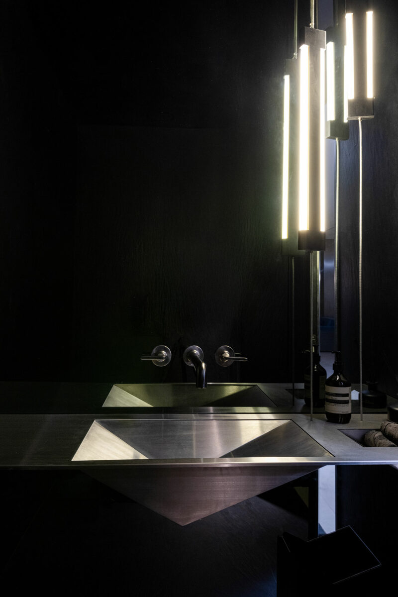 A modern bathroom with a geometric metal sink, wall-mounted faucet, dark walls, and vertical tube lights reflected in a mirror.