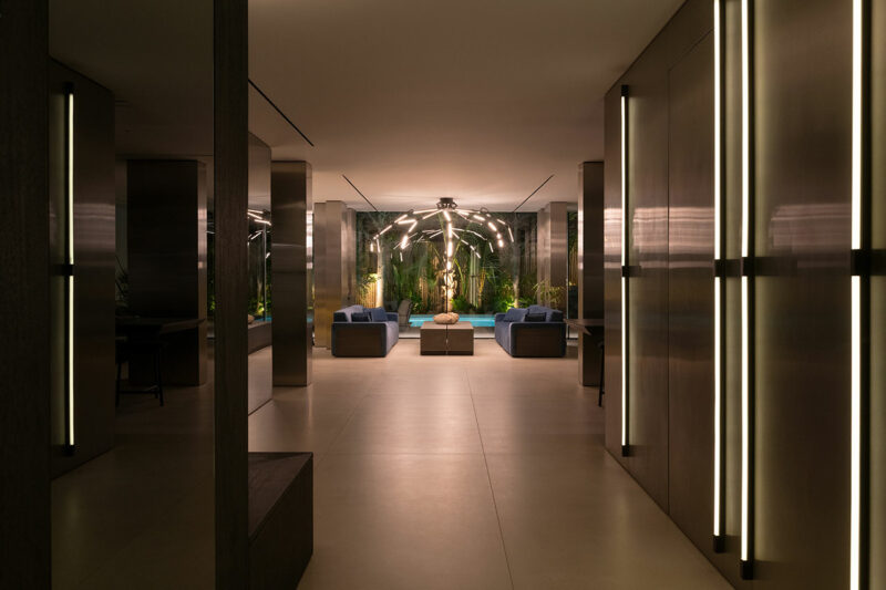 Modern lounge area with symmetrical lighting, dark walls, two blue sofas, and a decorative sculpture, seen from a spacious hallway with vertical light panels.