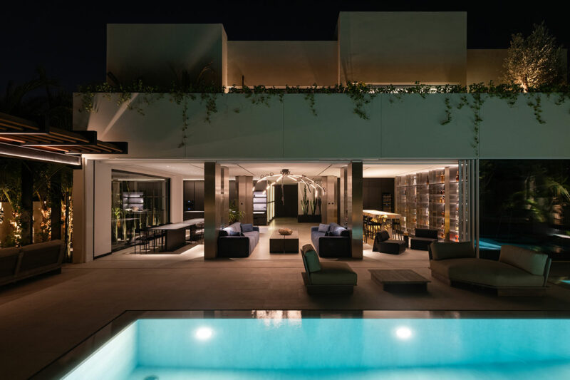 Modern house at night with illuminated pool, open living area, lounge seating, and indoor wine storage visible through glass walls.