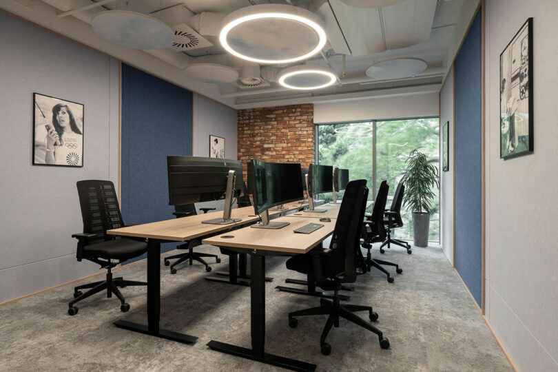 Modern BIT CREATIVE office space with six workstations, ergonomic chairs, multiple monitors, circular ceiling lights, brick accent wall, framed posters, and a large window overlooking greenery.