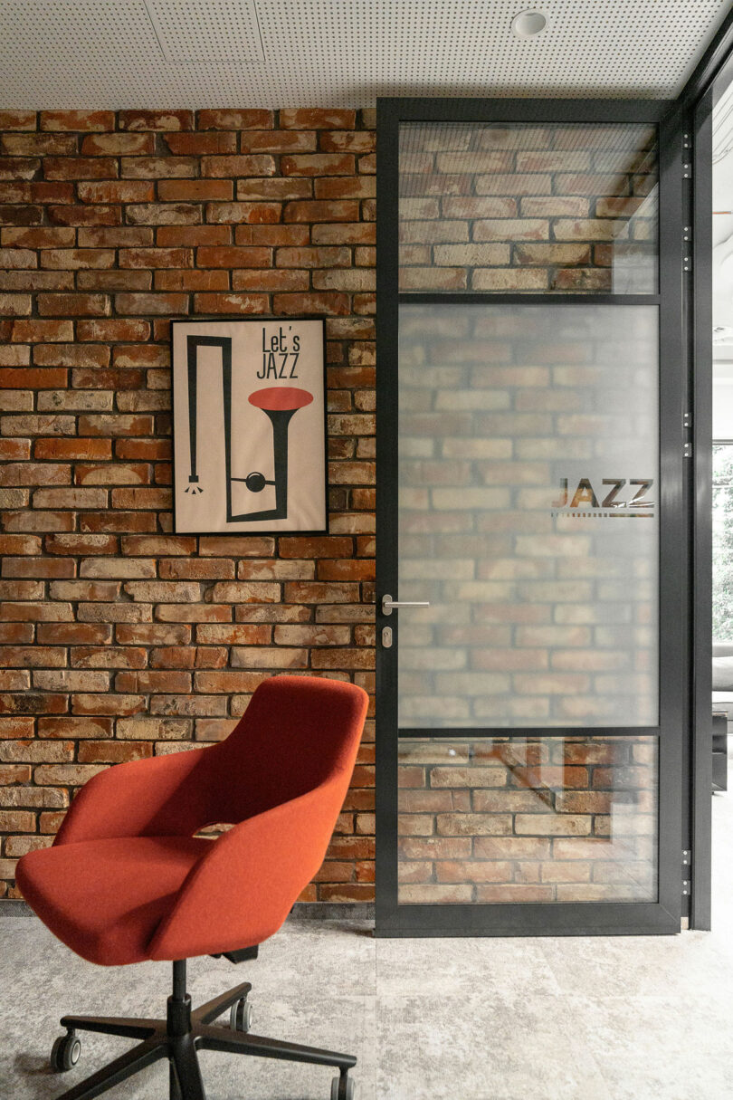 A modern office space by BIT CREATIVE features a red swivel chair, a brick wall, a framed