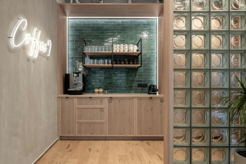 BIT CREATIVE small coffee station with a coffee machine, cups, glasses, and shelves against a green tile backsplash, next to a glass block wall and a “Coffee?” neon sign.