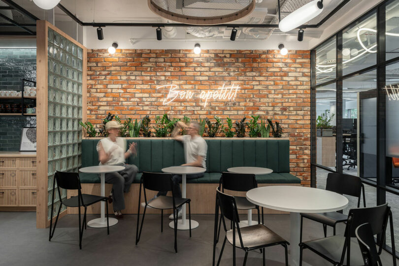 Modern office break area designed by BIT CREATIVE, featuring round tables, black chairs, green bench seating, a brick wall, neon
