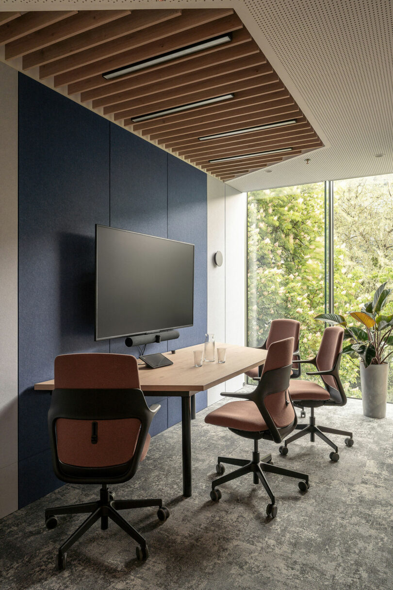 Modern BIT CREATIVE conference room with three office chairs around a wooden table, a large monitor on the wall, glass water bottles, and a large window revealing greenery outside.