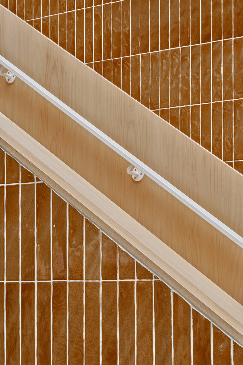 Close-up of a light wood handrail set diagonally against a wall covered with glossy, rectangular brown tiles arranged in a vertical grid pattern.