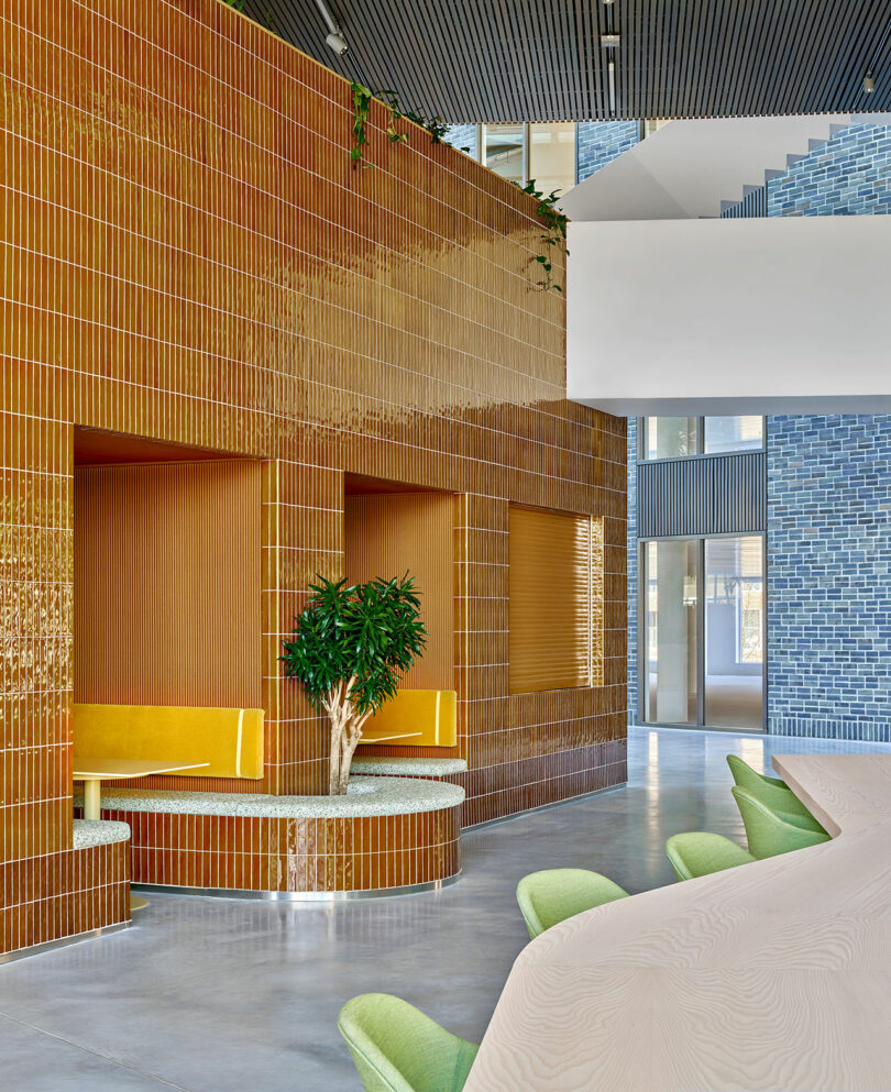 Modern interior with mustard yellow tiled booths, a small potted tree, mint green chairs, a light wood table, and blue tiled walls in the background.
