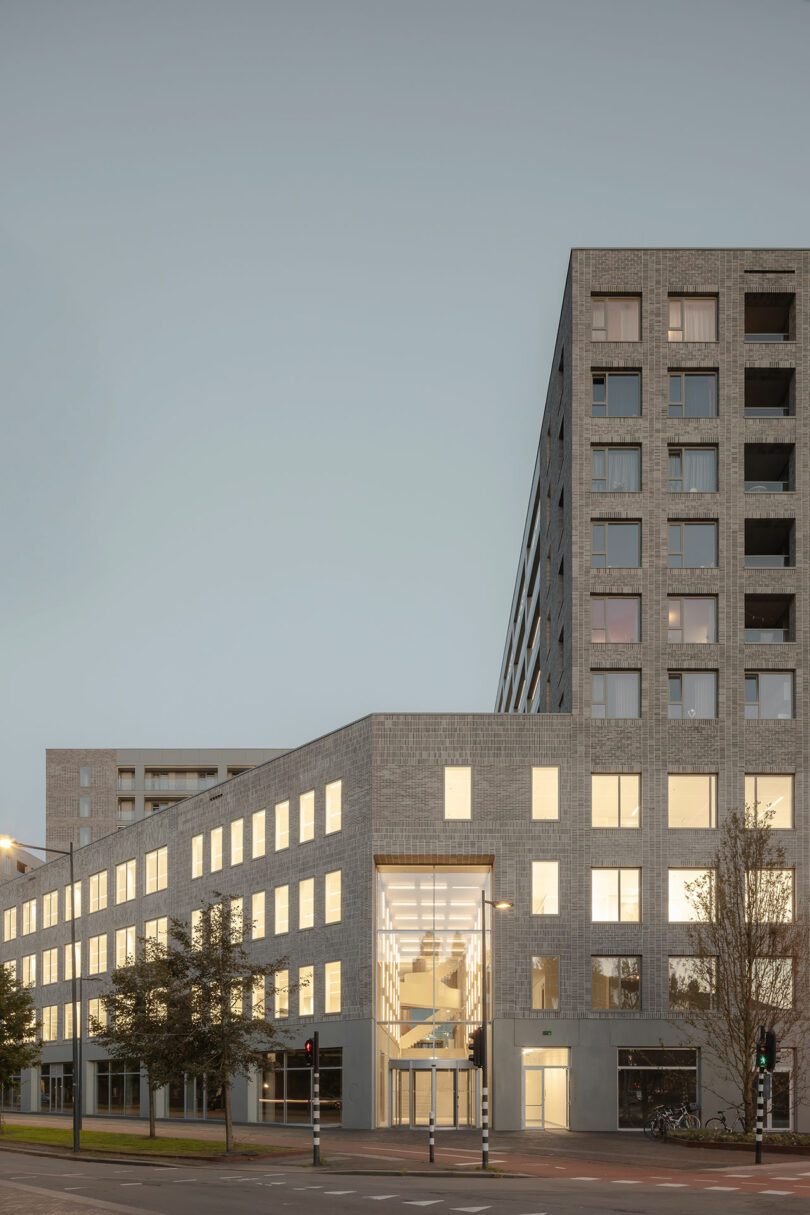Modern multi-story building with large windows, some illuminated, situated at a street intersection with trees and traffic lights in the foreground.