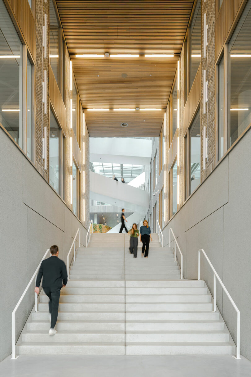 People walk up and down a wide, white staircase inside a modern building with high ceilings, large windows, and linear ceiling lights.