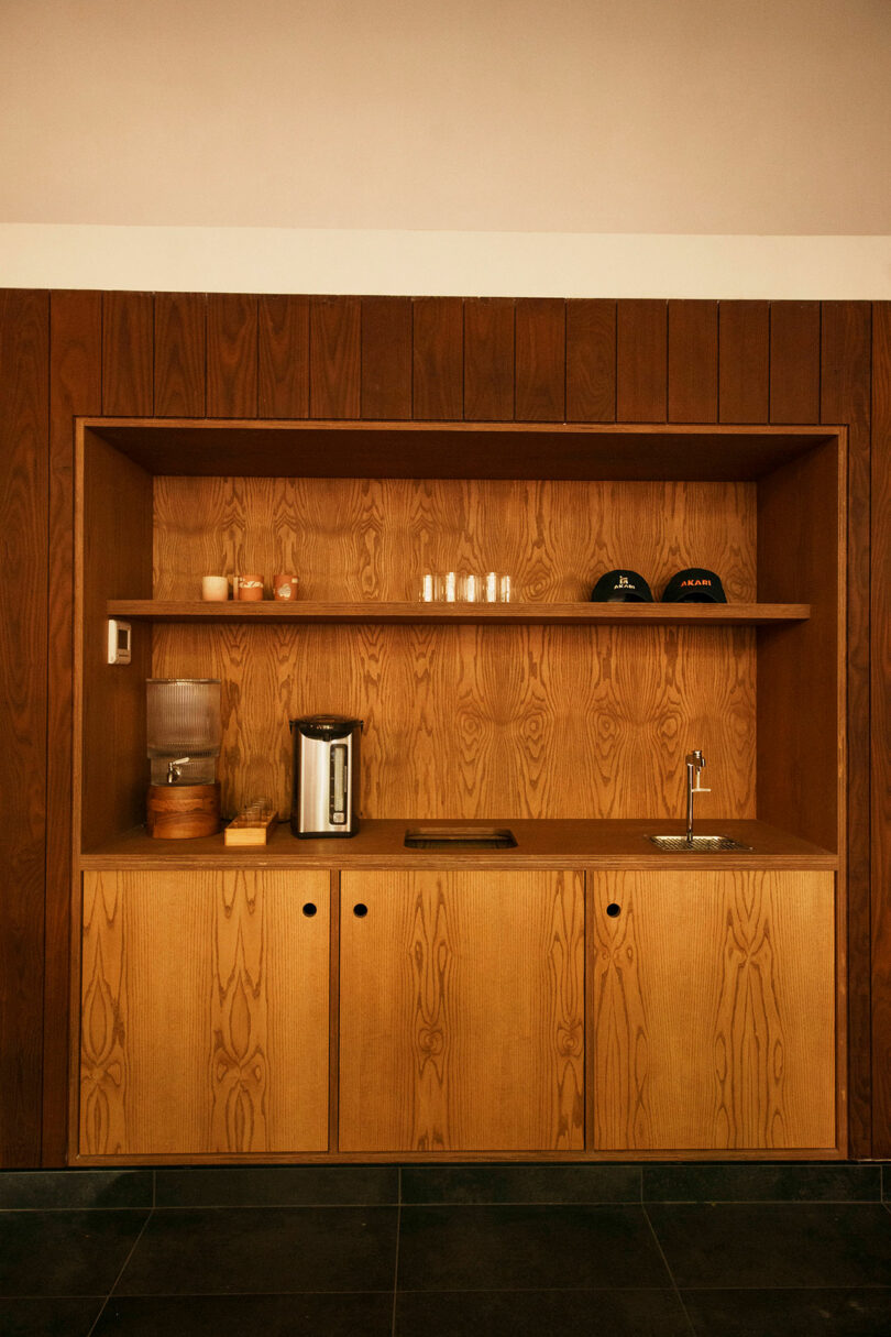 A wooden kitchenette with a countertop, sink, electric kettle, cups, water dispenser, and a shelf holding mugs, glasses, bottles, and two black caps.