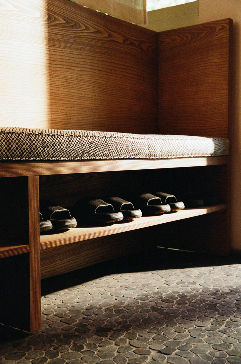 Wooden bench with a patterned cushion on top and four pairs of black slippers neatly arranged on a lower shelf, set on a stone tile floor.