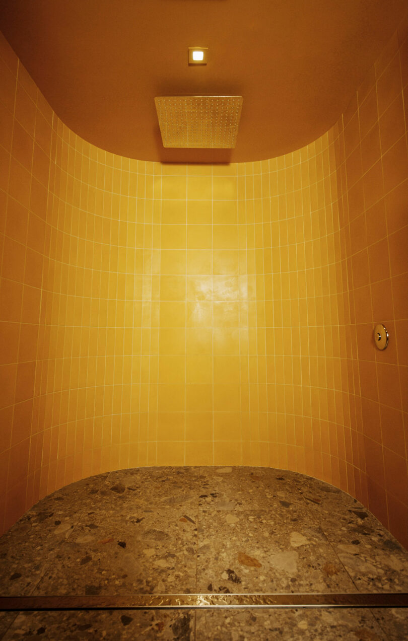 Yellow-tiled shower with a square ceiling-mounted showerhead, a round control knob on the right wall, and a speckled stone floor.