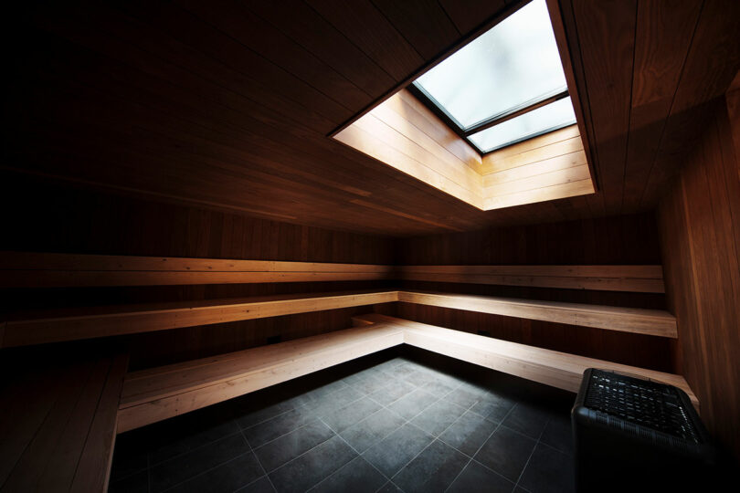 A modern wooden sauna room with tiered benches, a tiled floor, a heater in the corner, and a large skylight letting in natural light.