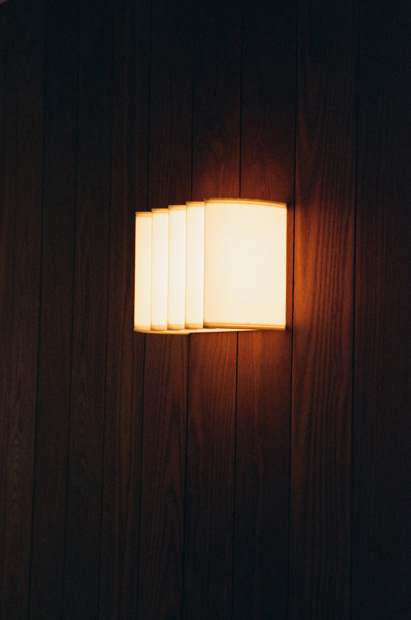 A modern wall sconce with four rectangular lights emits a warm glow against a dark wooden panel wall.