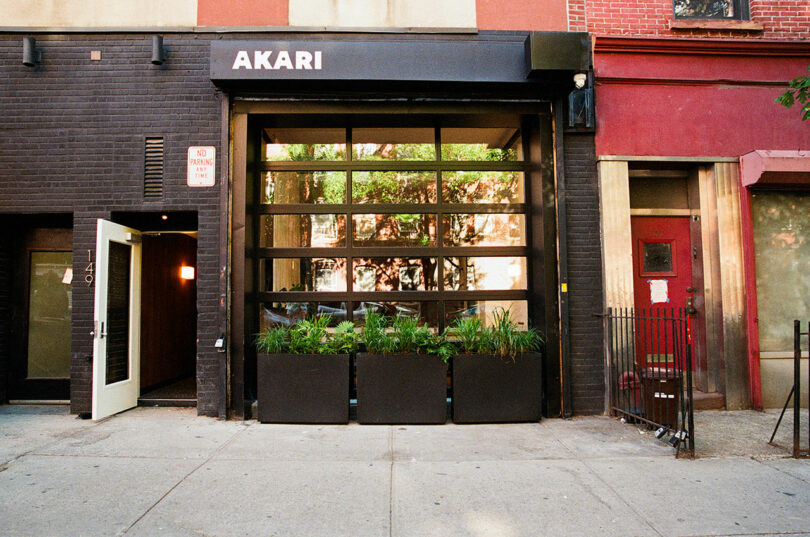 Street view of the Akari restaurant entrance with glass garage-style door, black planters with greenery, and an open side door at 149, on a city sidewalk.